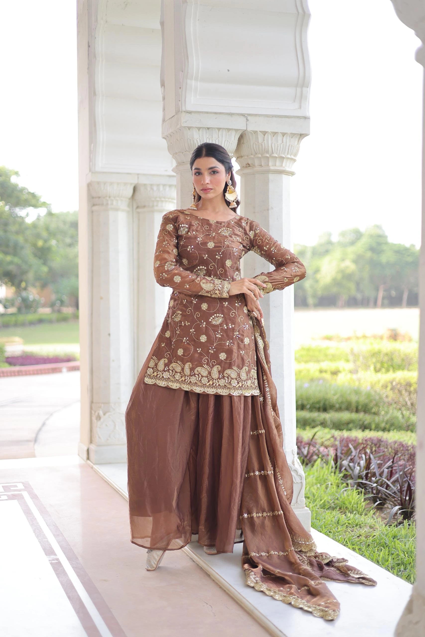 Woman in a brown traditional outfit standing in front of a white architectural structure with greenery in the background.