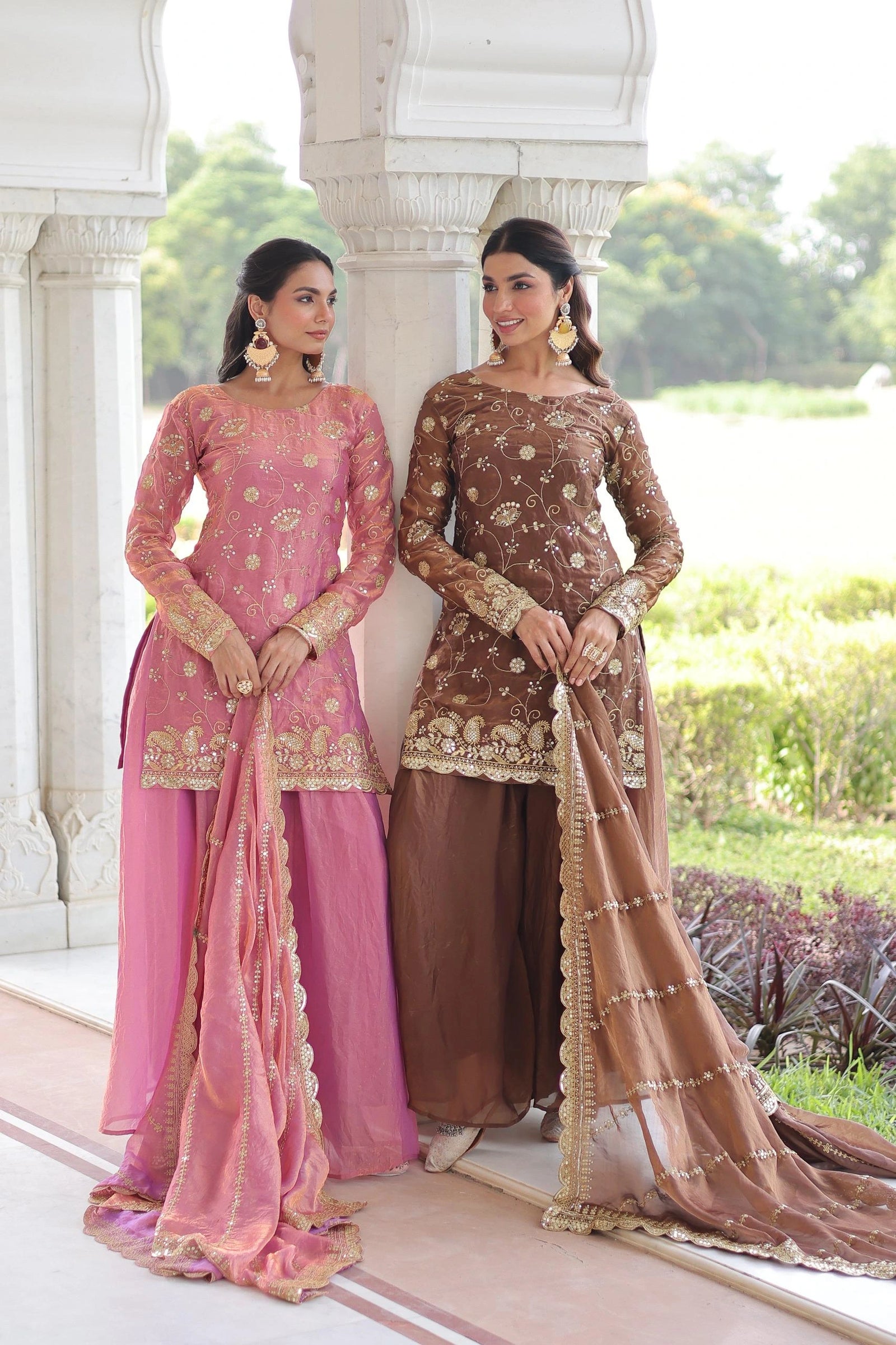 Two women in traditional pink and brown outfits standing outdoors with a scenic background.