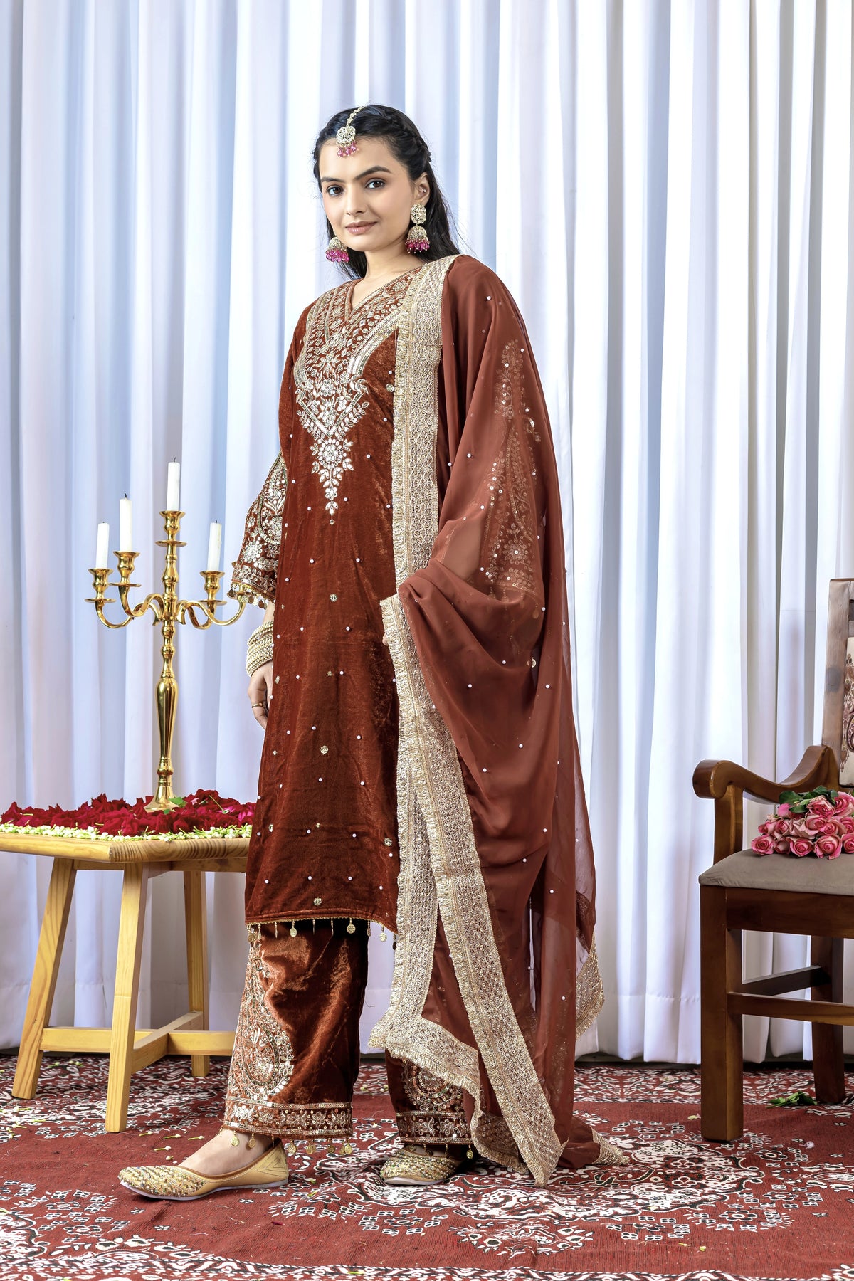 Woman in a traditional brown and beige embroidered outfit standing in a decorated room with white curtains and a table.
