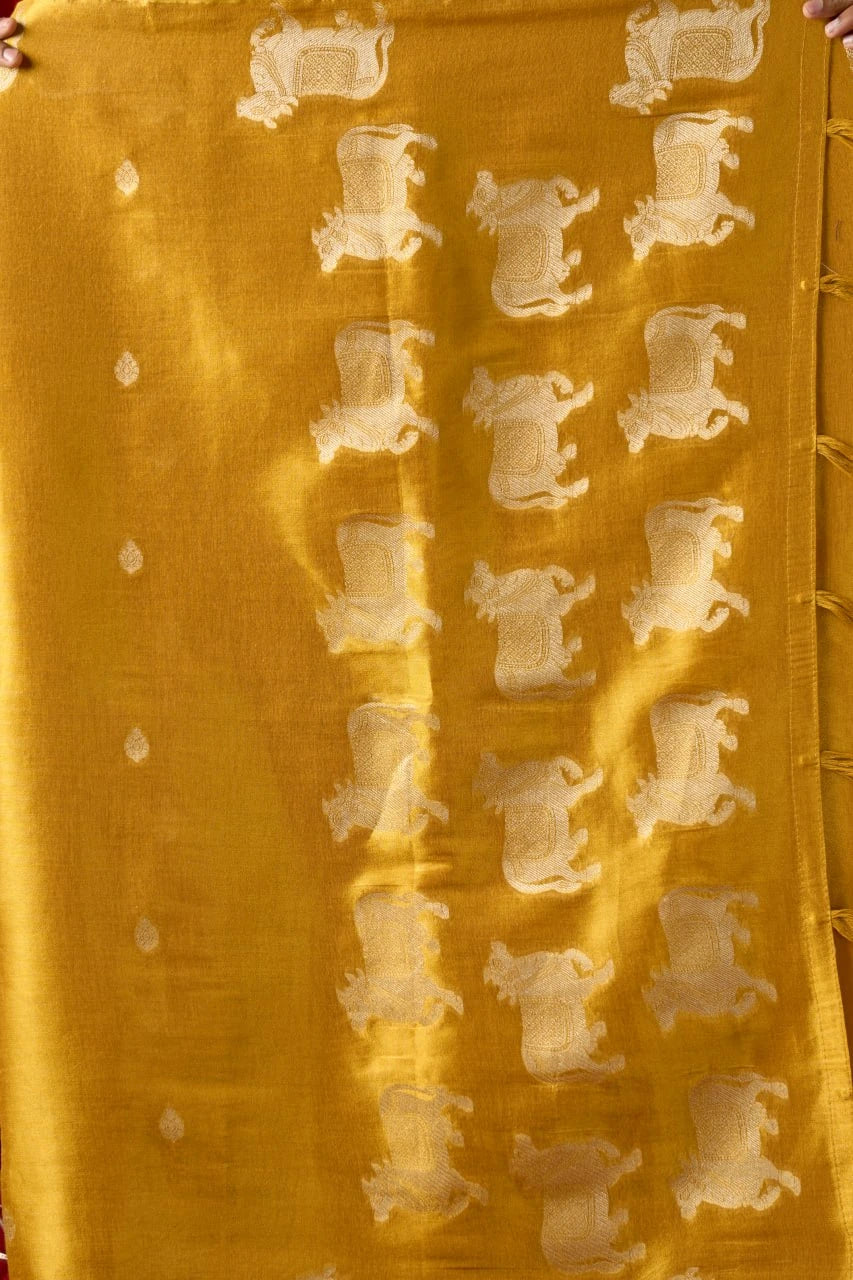 A close-up, overhead photograph of a mustard-yellow textured textile panel. It features repeating vertical columns of intricately woven, pale-gold zari thread motifs of galloping horses. To the left is a column of smaller circular zari motifs. The right edge has a border with knotted tassel fringe, and hands are visible at the very top corners holding the fabric.