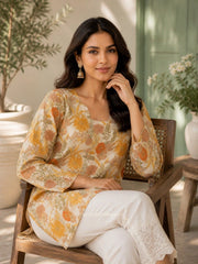 Woman sitting on a wooden chair wearing a floral top and white pants in an indoor setting with plants.
