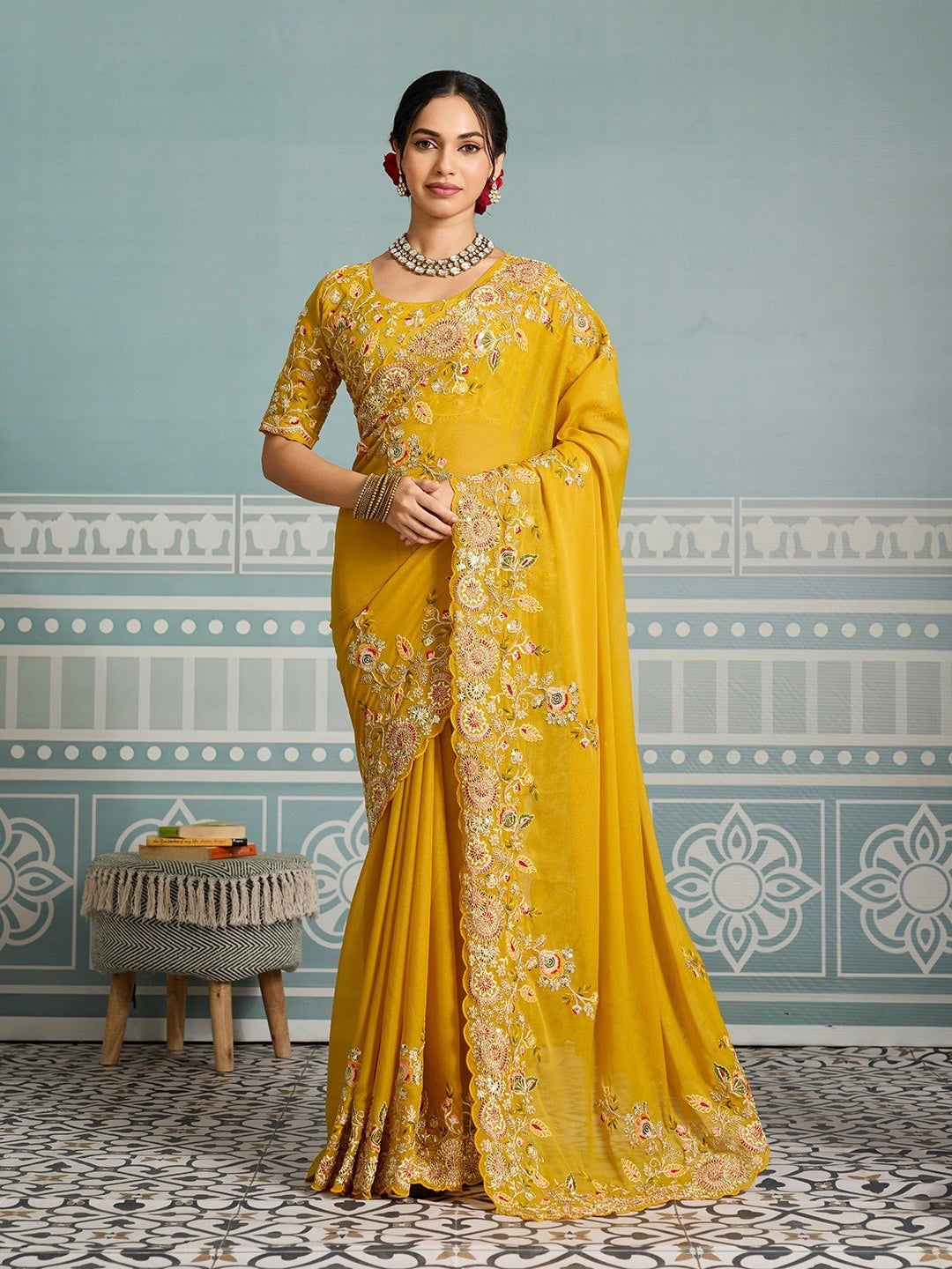 Woman wearing a yellow saree with intricate patterns in a room with decorative wallpaper.