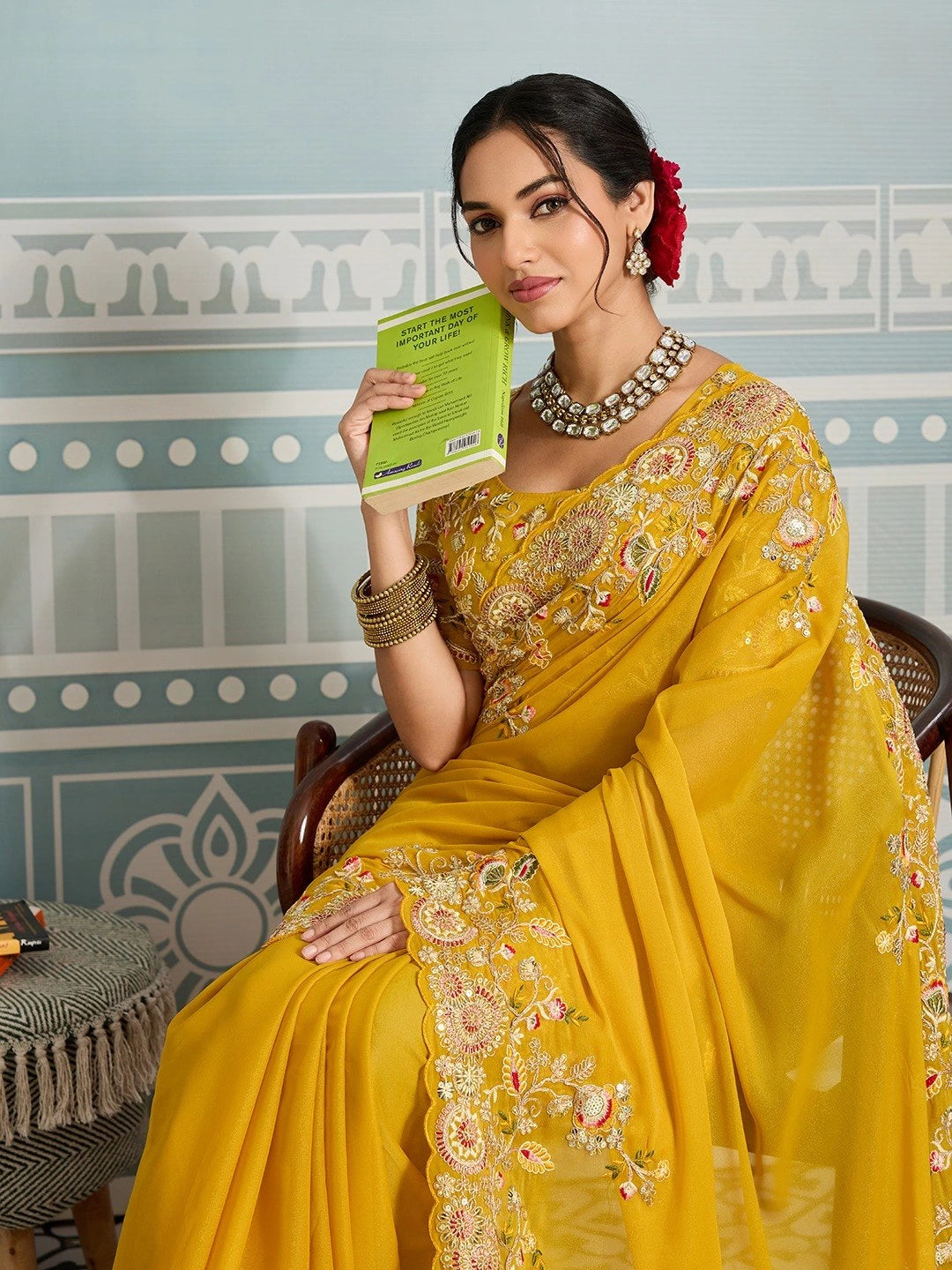 Woman in a yellow saree holding a book against a decorative wall.