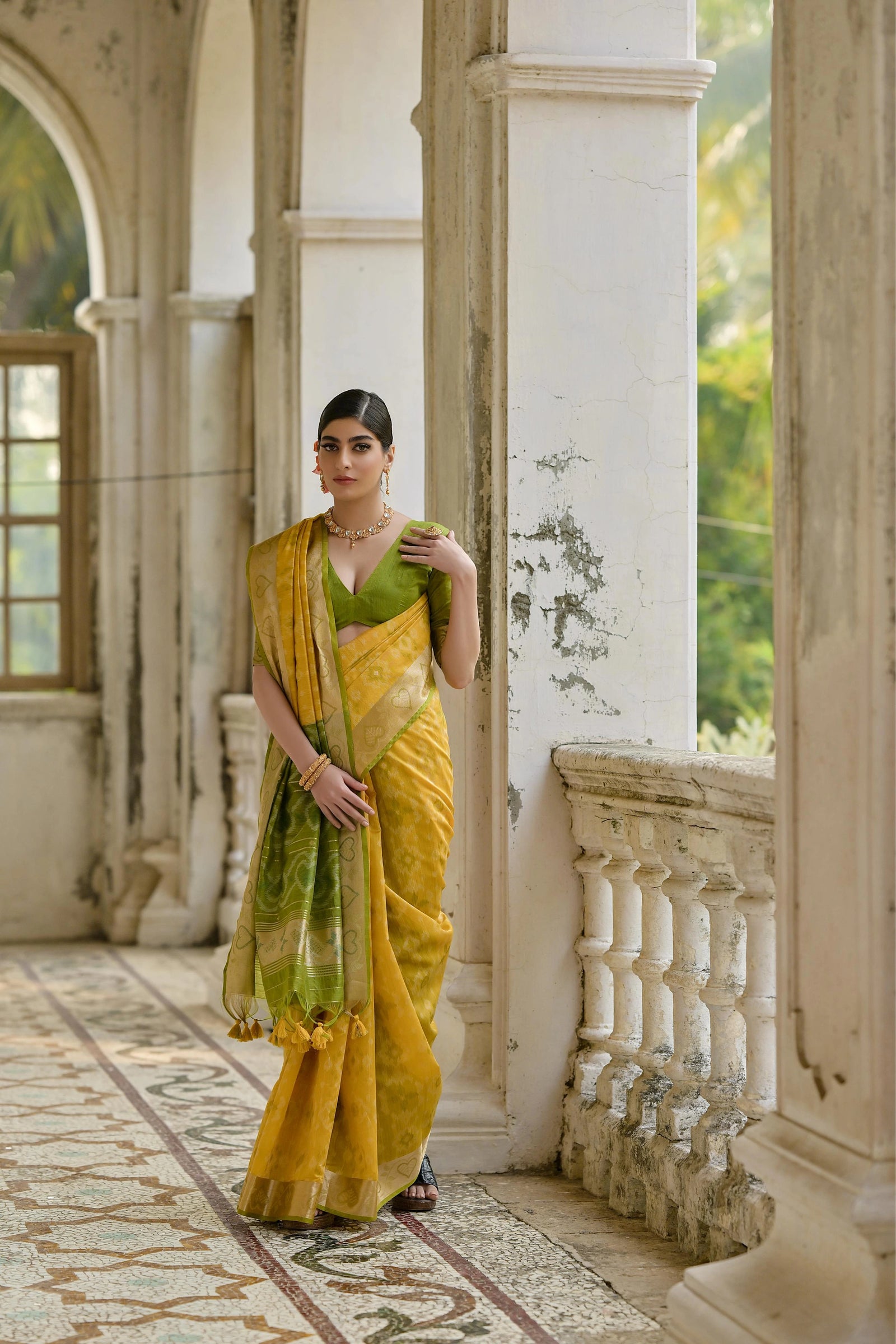 Woman in a yellow and green saree standing in an elegant indoor setting.