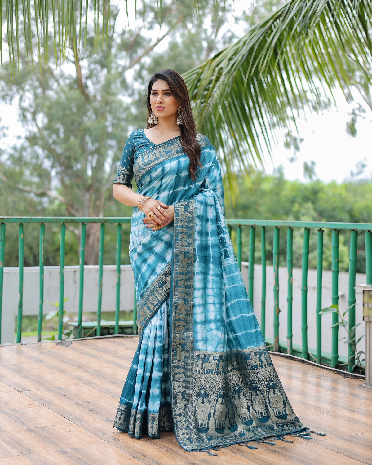 Woman wearing a blue and green saree on a wooden deck with greenery in the background
