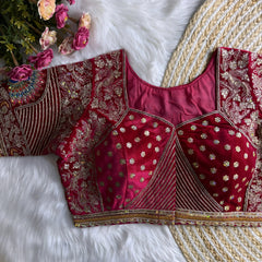 Traditional maroon silk blouse featuring intricate gold zardosi embroidery with floral and peacock motifs, sequin embellishments, geometric panel designs, and decorative border, displayed flat on a white fur surface with a braided natural mat and pink artificial flowers in the corner.