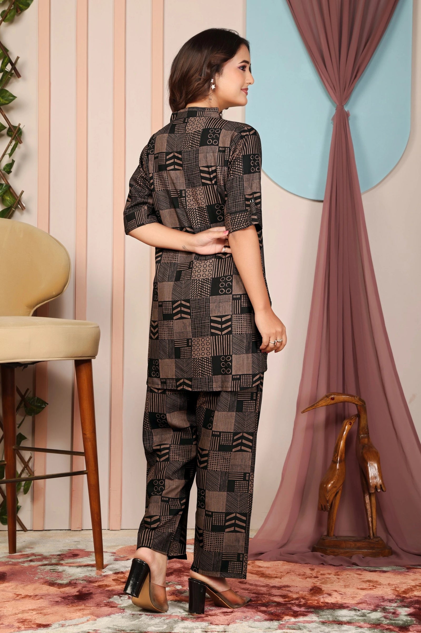 Woman wearing a patterned outfit standing in a room with a chair and decorative elements.
