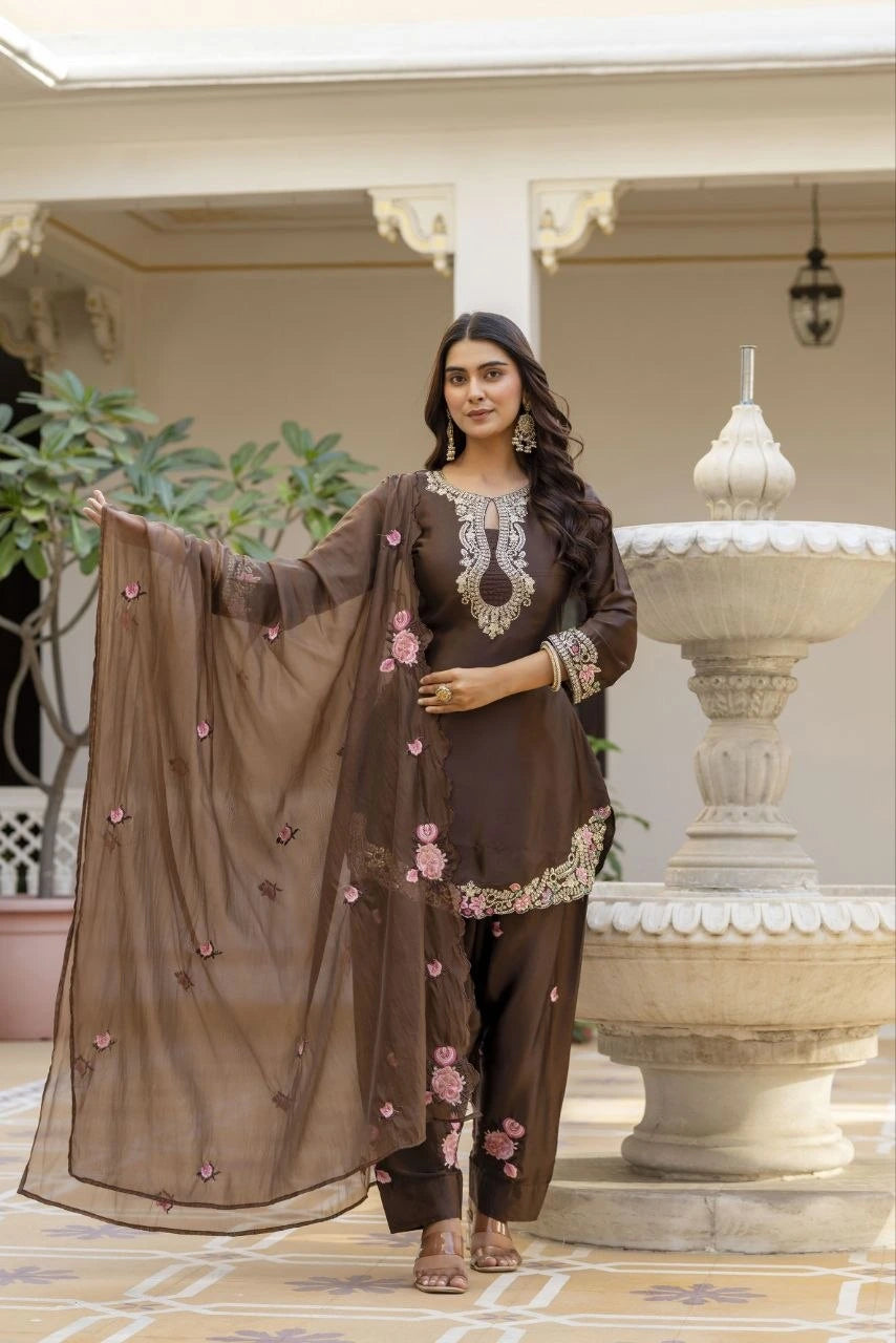 Woman in a brown traditional outfit with floral details standing in front of a decorative fountain.