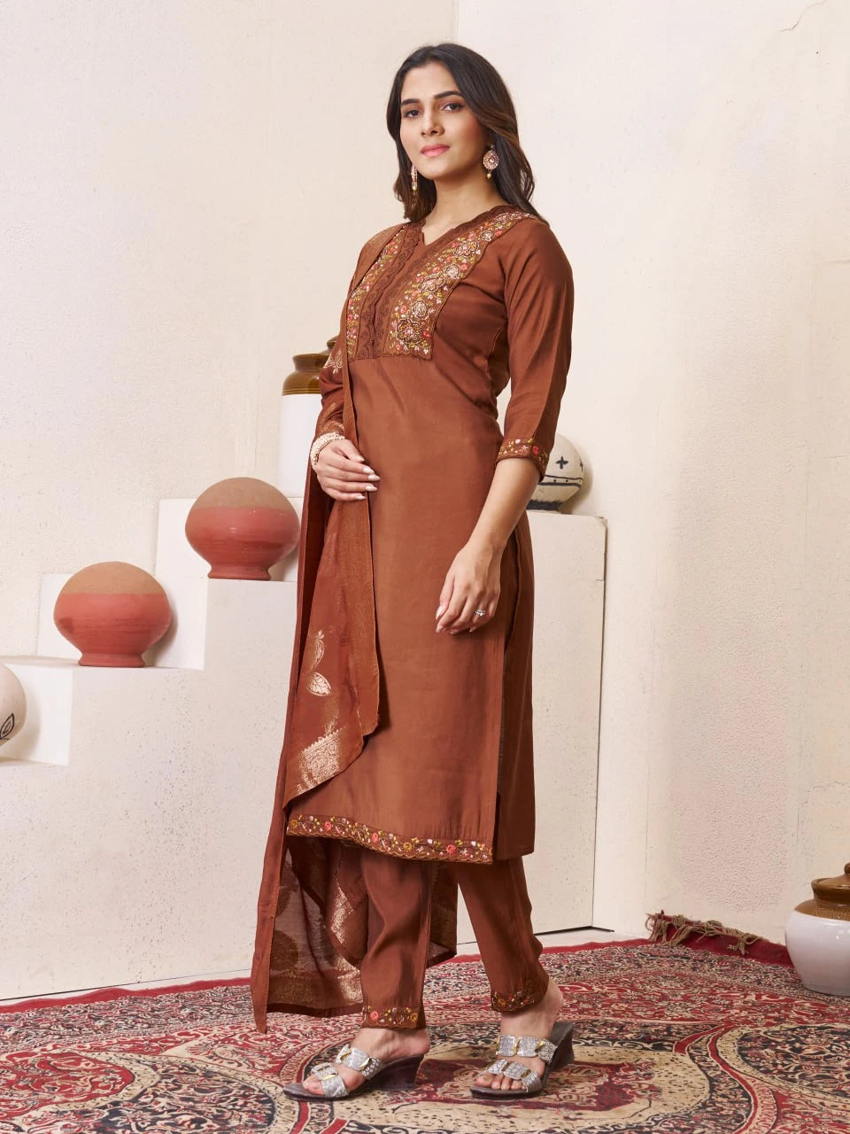 Woman wearing a brown traditional outfit with intricate designs in a room with decorative elements.