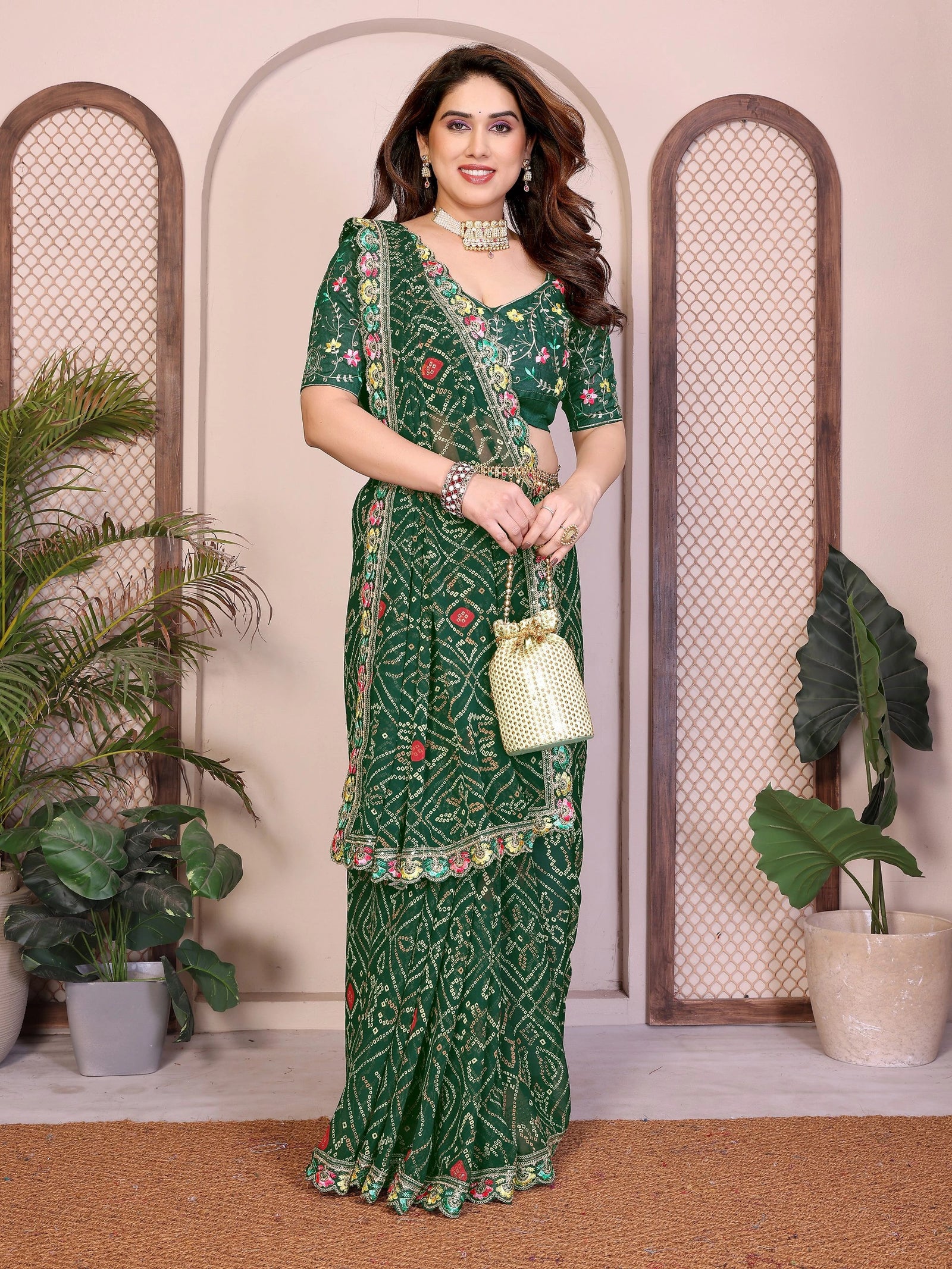 Woman in a green saree with floral patterns standing indoors with decorative plants.