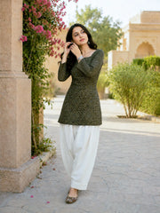 Woman in a green patterned top and white pants standing outdoors near a building with greenery.