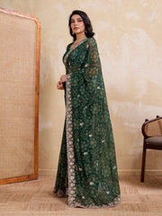 Woman wearing a green embroidered saree in a room with a textured wall and chair.