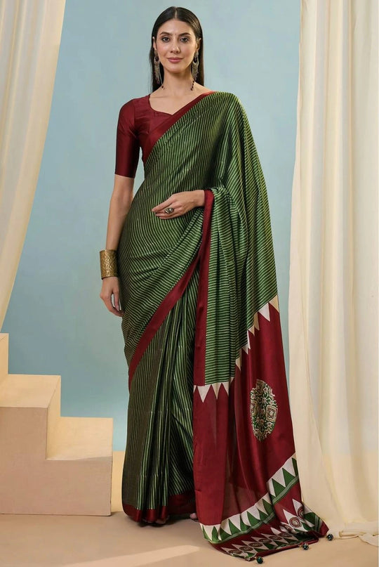 Woman wearing a green and red saree with a decorative pattern, standing against a light blue wall.