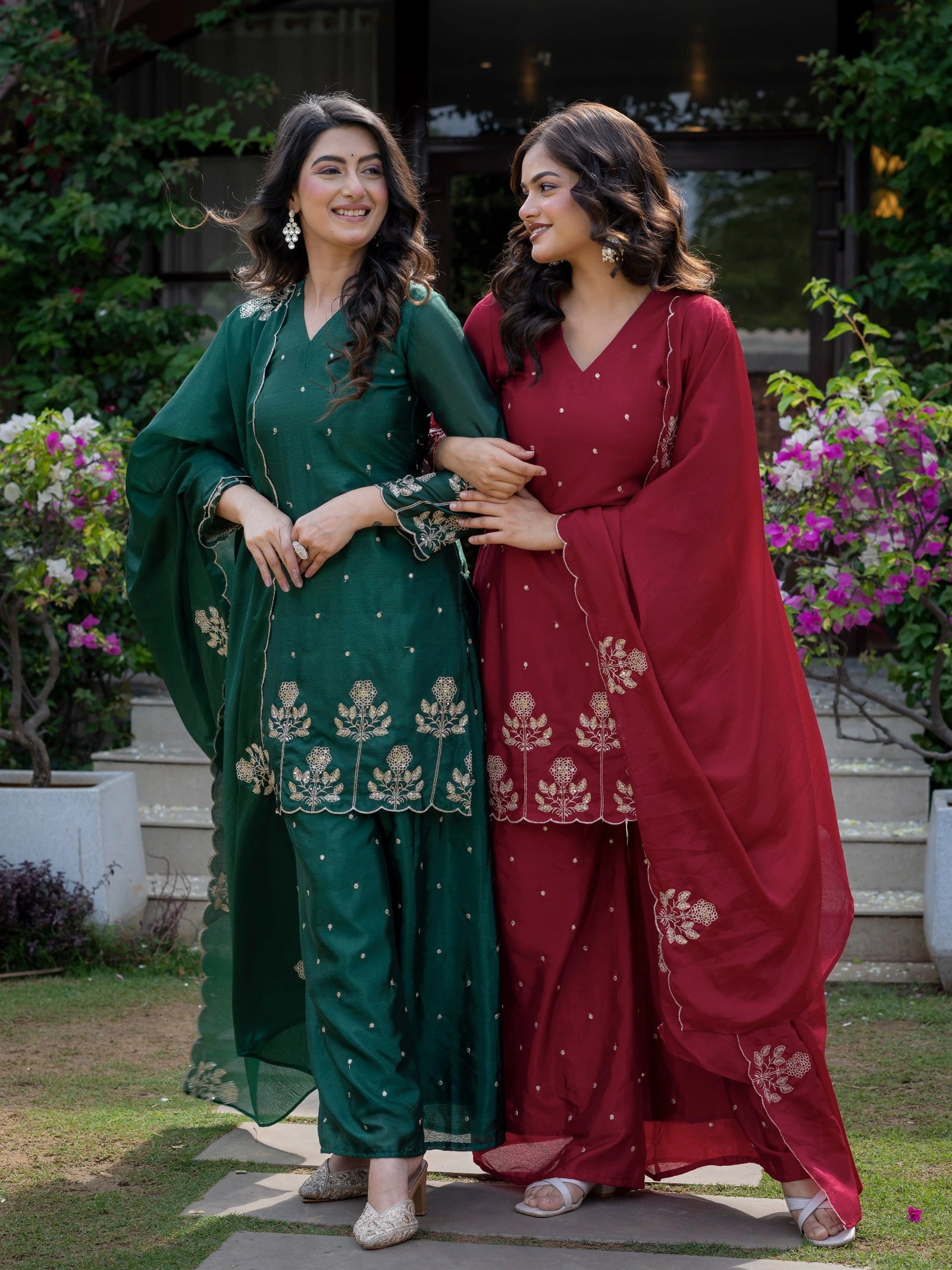 Two women in traditional green and red outfits standing outdoors with flowers and plants in the background.