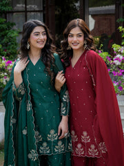 Two women in traditional green and red outfits standing outdoors with flowers in the background