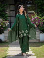 Woman in a green traditional outfit standing outdoors with plants and flowers in the background
