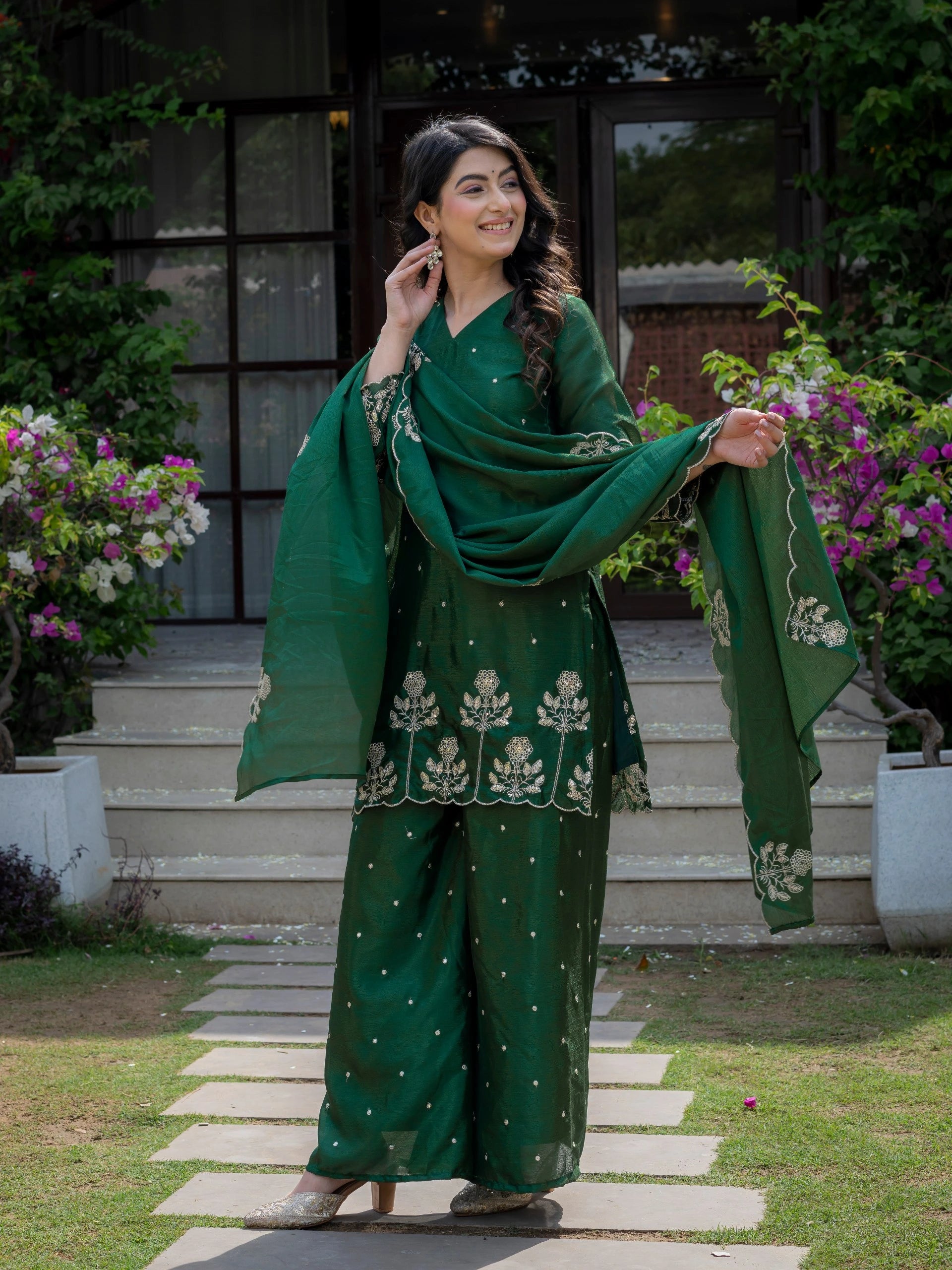 Woman in a green traditional outfit standing outdoors with flowers and steps in the background