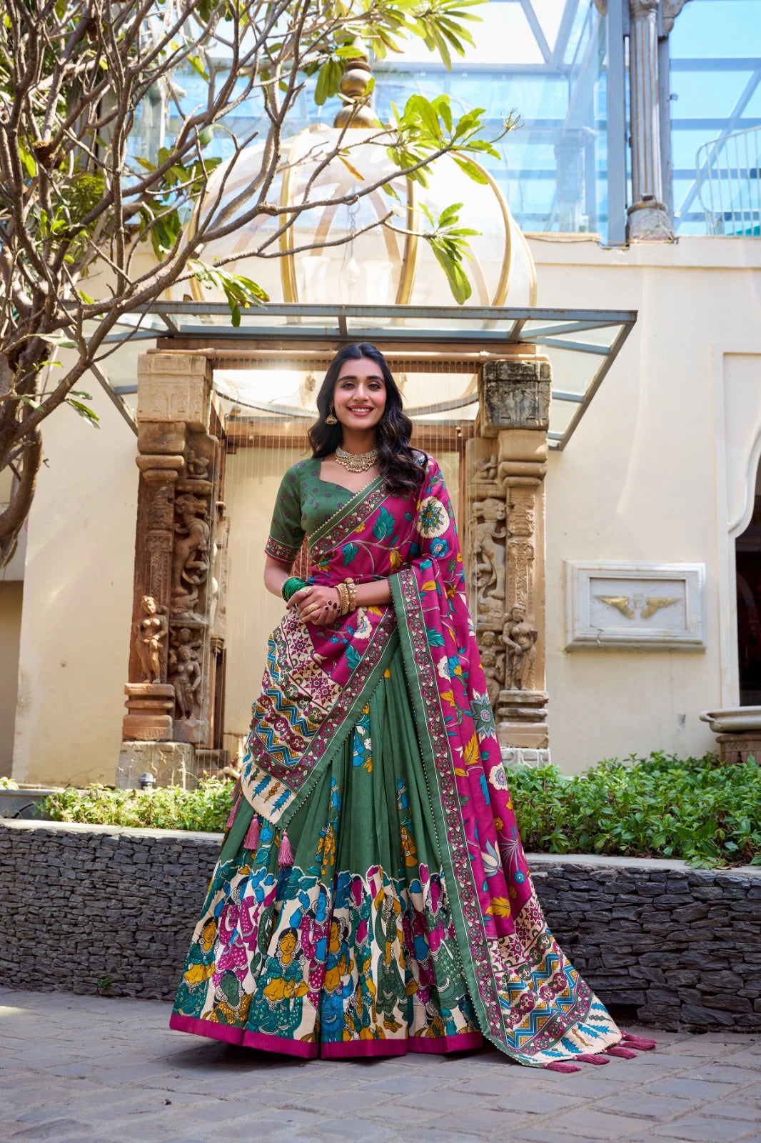 Woman in a colorful saree standing in an outdoor setting with architectural elements.