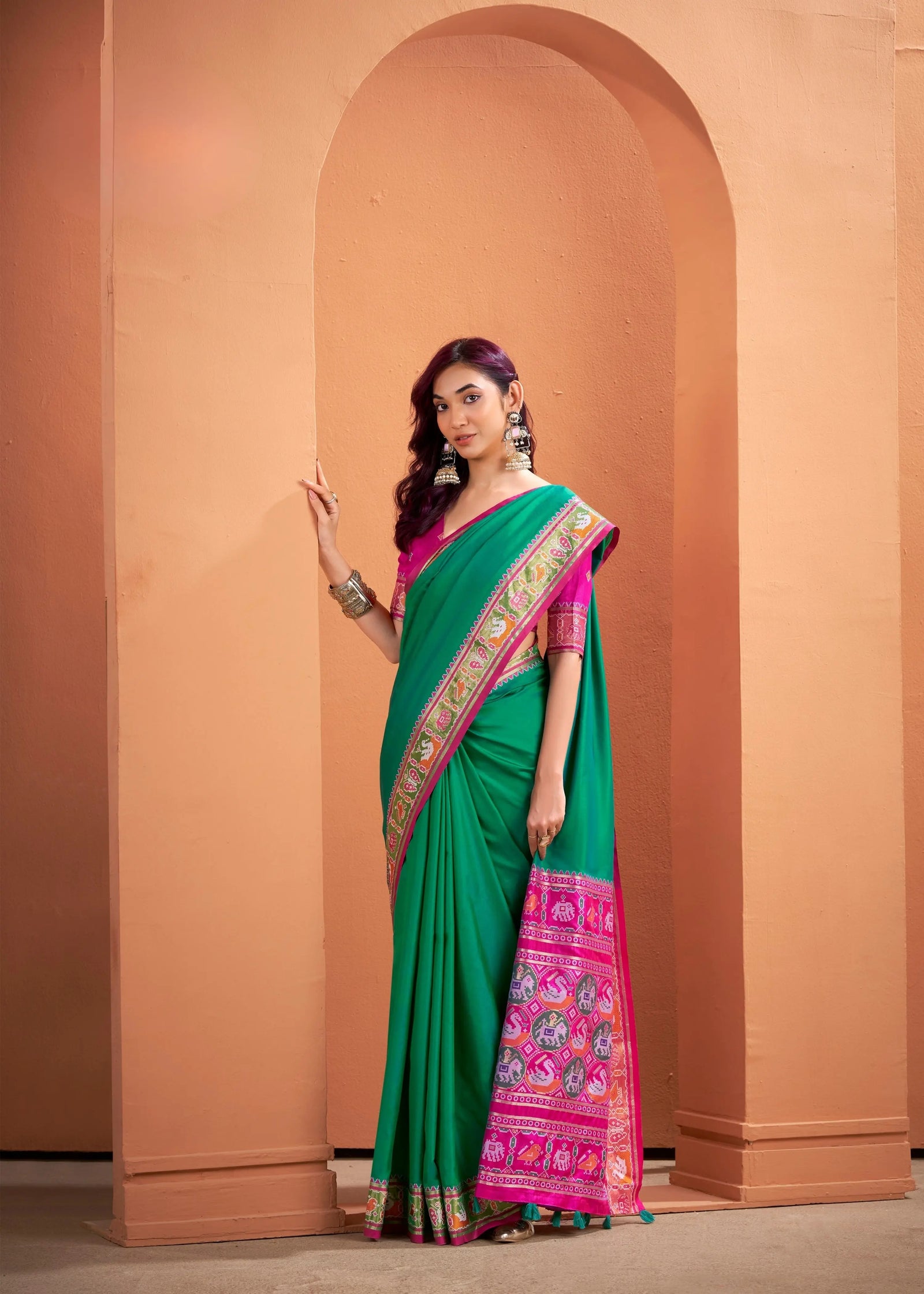 Woman wearing a green saree with pink border against a peach-colored wall.