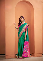 Woman wearing a green saree with pink border against a peach-colored wall.