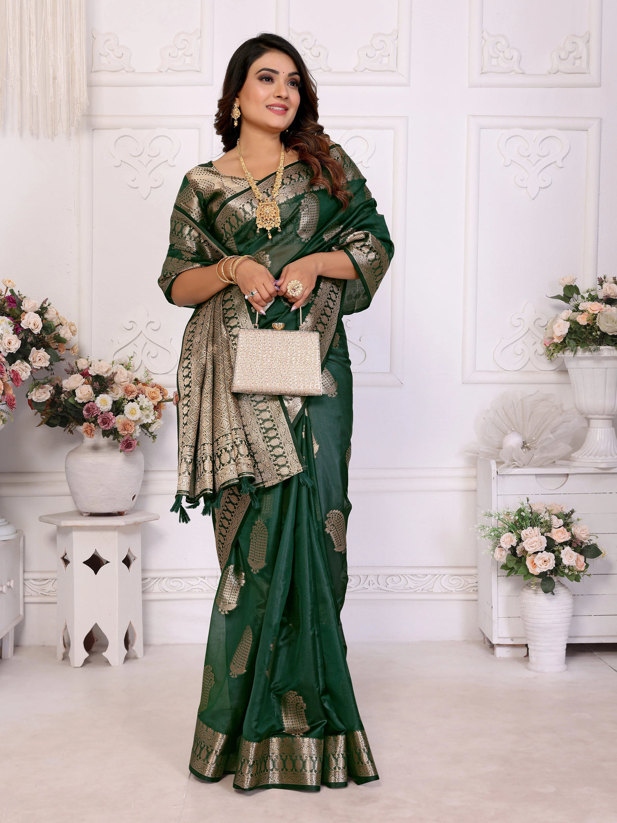 Woman in a green saree with gold patterns standing in a decorated room with flowers and decorative items.