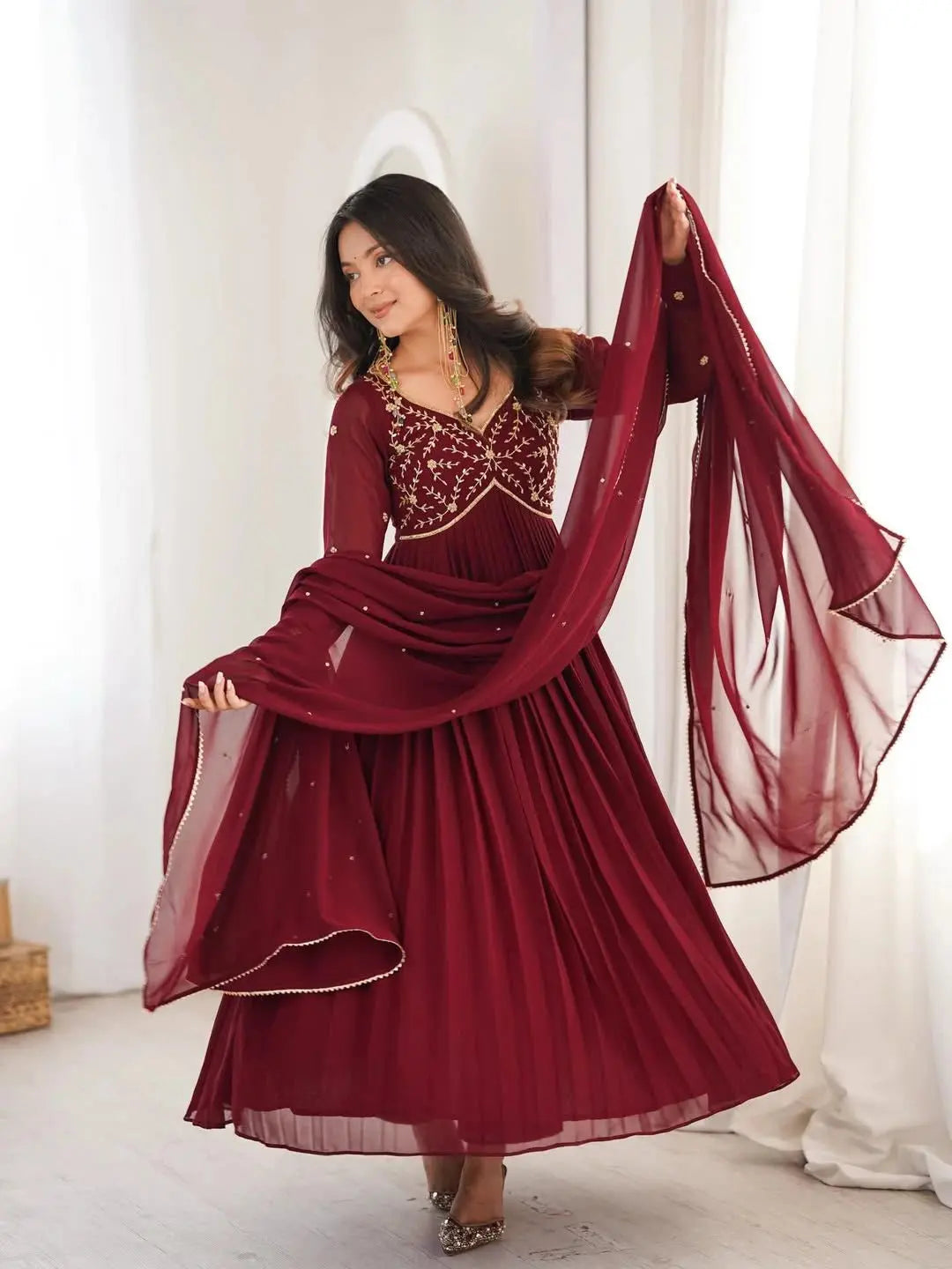 Woman in a burgundy evening gown with sheer overlay in a bright room.