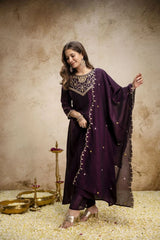 Woman in a dark purple traditional outfit with gold embellishments against a textured beige wall.