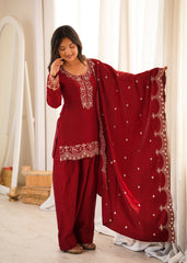 Woman in a red traditional outfit with white embroidery standing indoors.