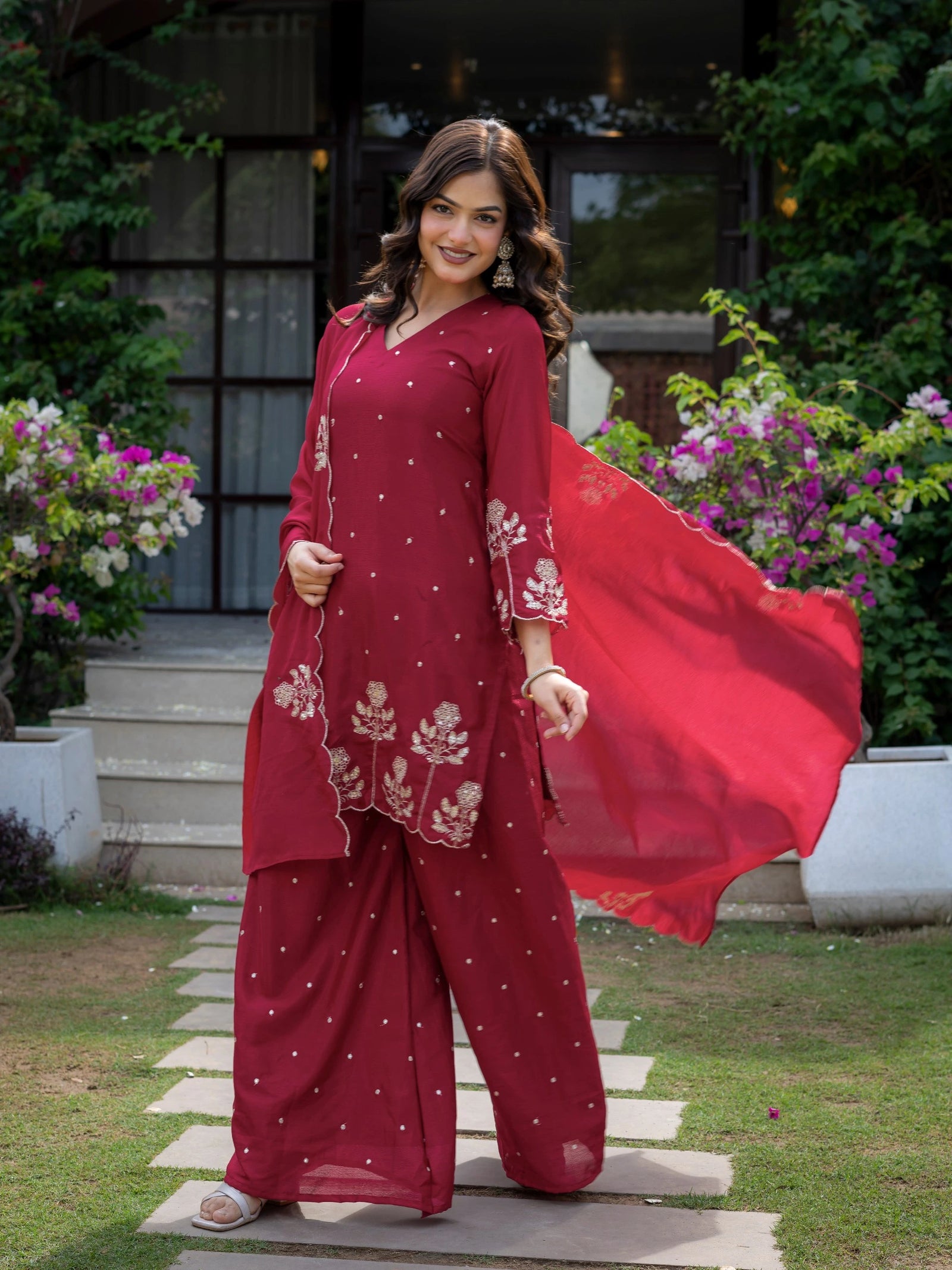 Woman in a red traditional outfit standing outdoors with flowers and greenery around.