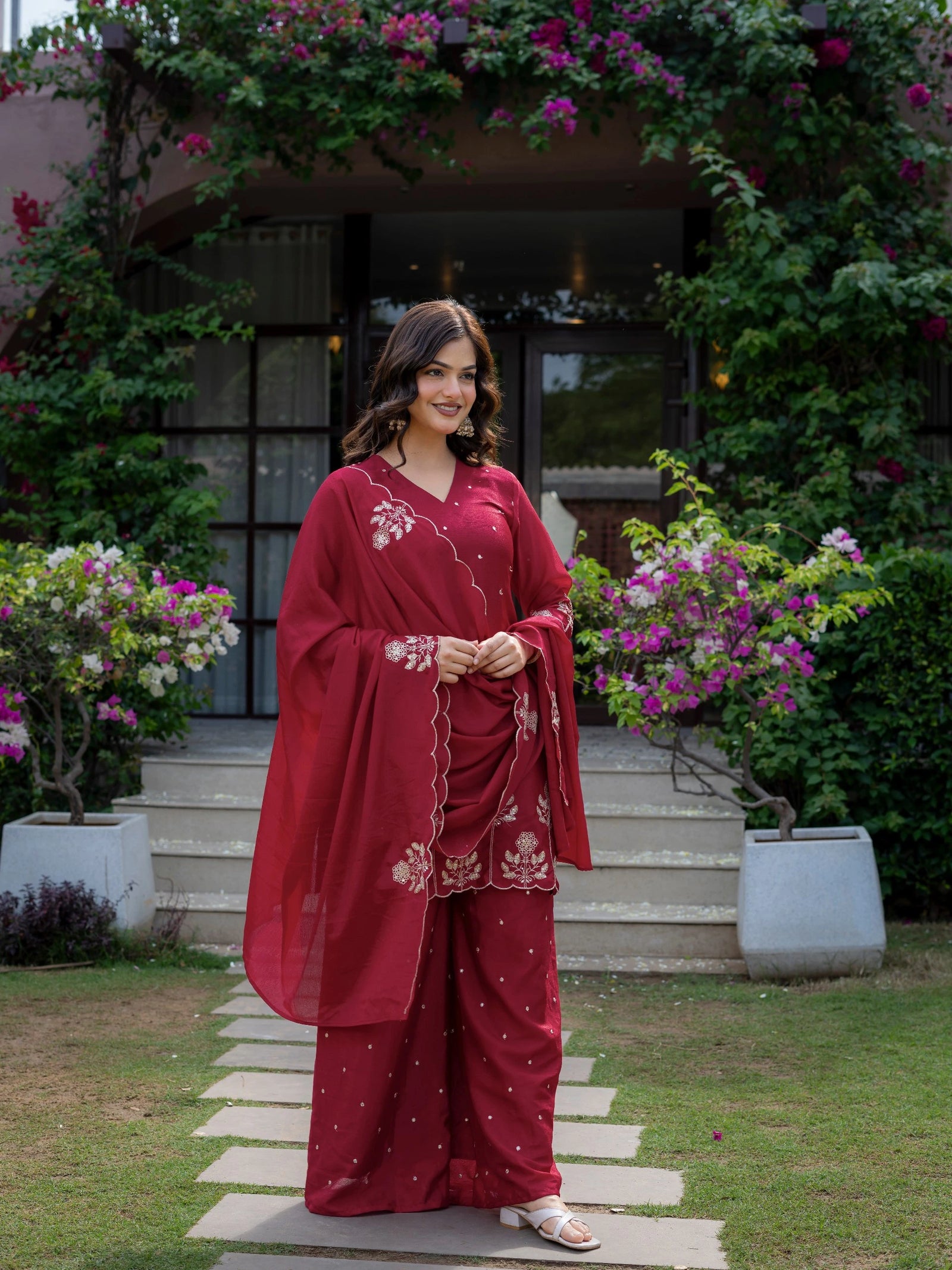 Woman in a red traditional outfit standing on a stone path with flowers and plants around.