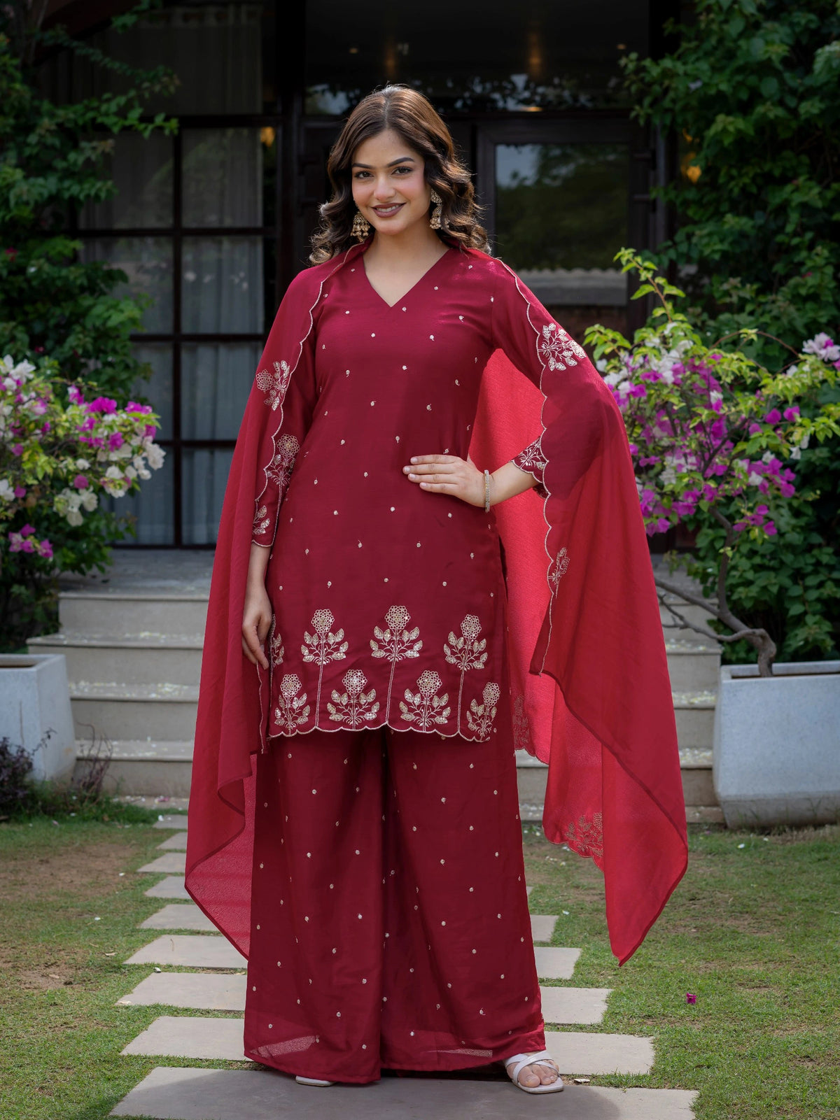 Woman in a red traditional outfit standing outdoors with flowers and steps in the background