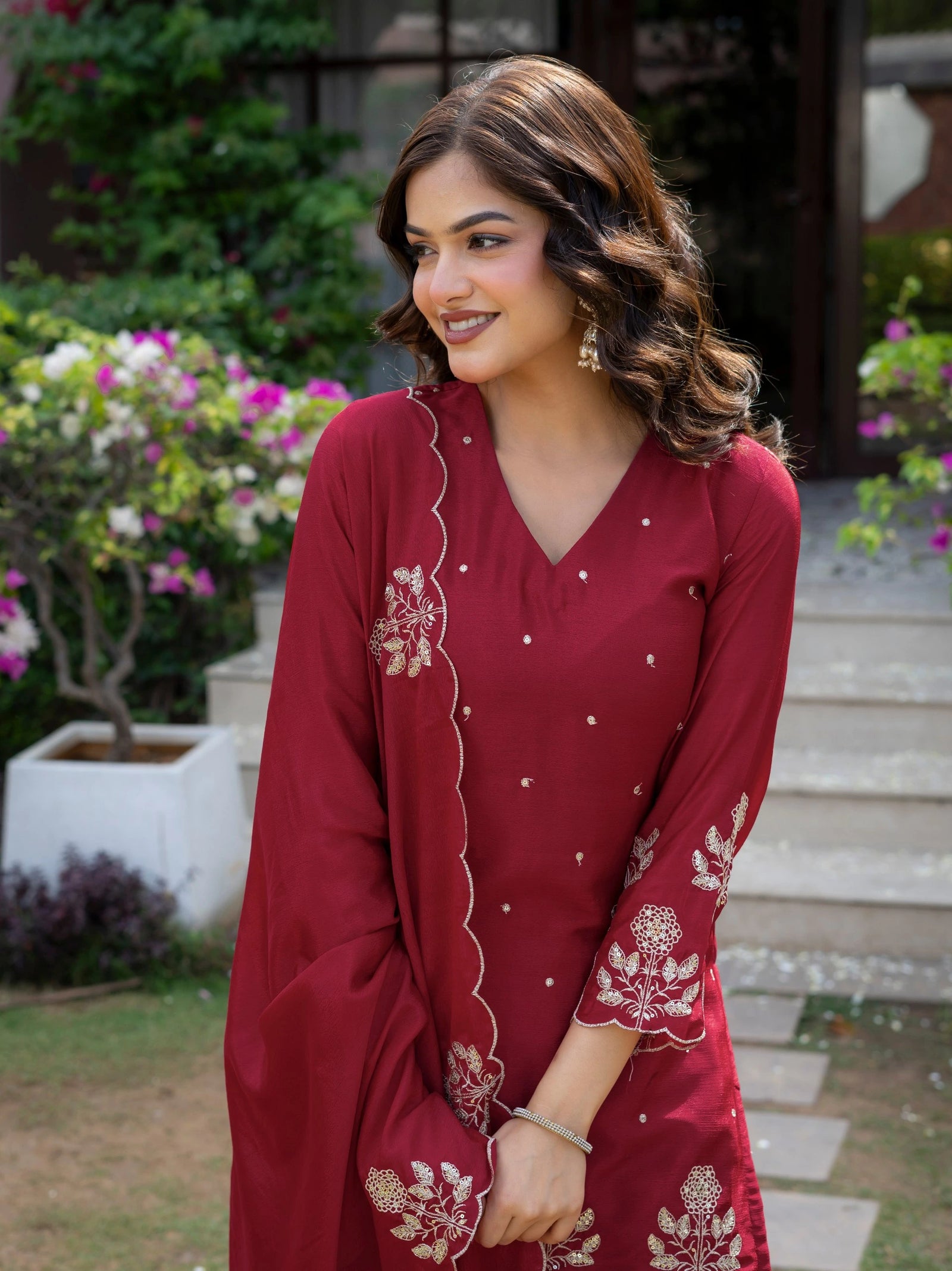 Woman wearing a red embroidered outfit in an outdoor setting with plants and flowers.