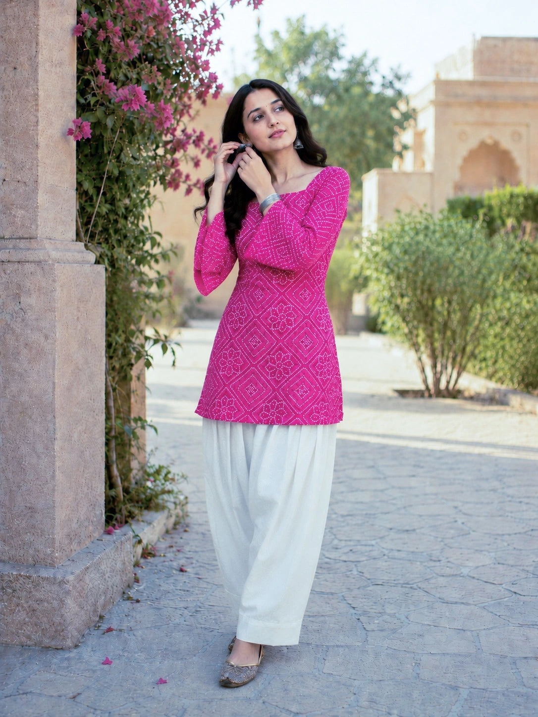 Woman in a pink top and white pants standing outdoors with greenery and architecture in the background