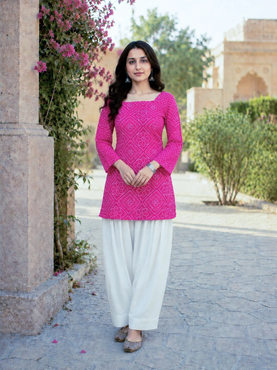 Woman wearing a bright pink top and white pants standing outdoors with greenery and architecture in the background.