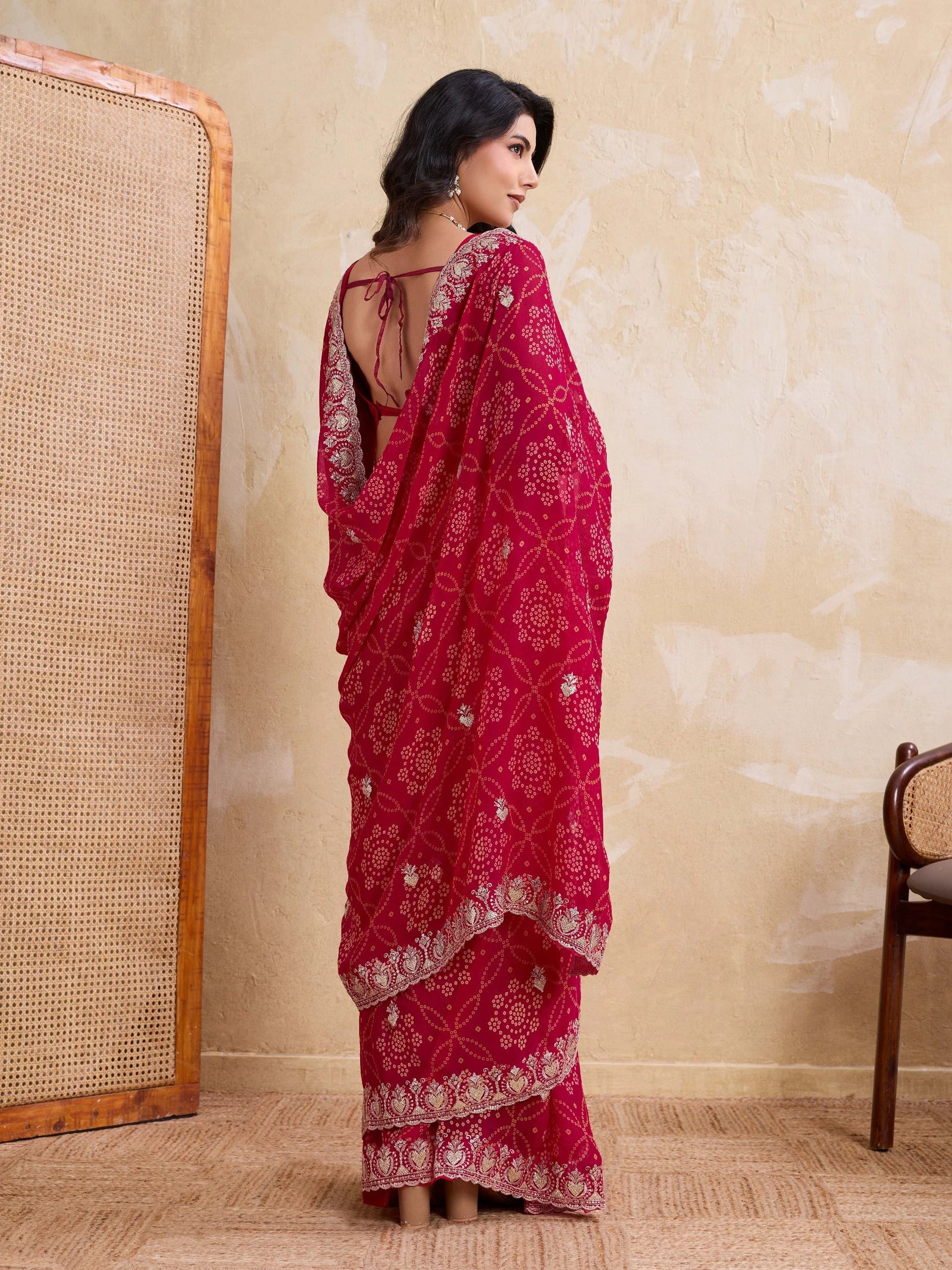 Woman wearing a red saree with white patterns in a room with beige walls and wooden furniture.