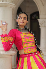 Woman in a traditional pink and yellow outfit with intricate patterns, standing against an architectural background.
