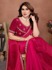 Woman in a pink saree with intricate designs sitting on a chair against a brown background.