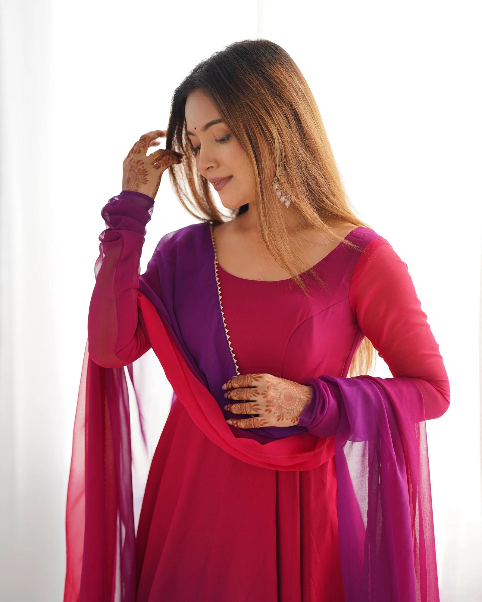 A medium shot of a woman in a magenta and purple Anarkali-style suit with a matching dupatta, looking down thoughtfully while touching her hair. Her hands are decorated with intricate henna patterns, and she wears ornate silver drop earrings.
