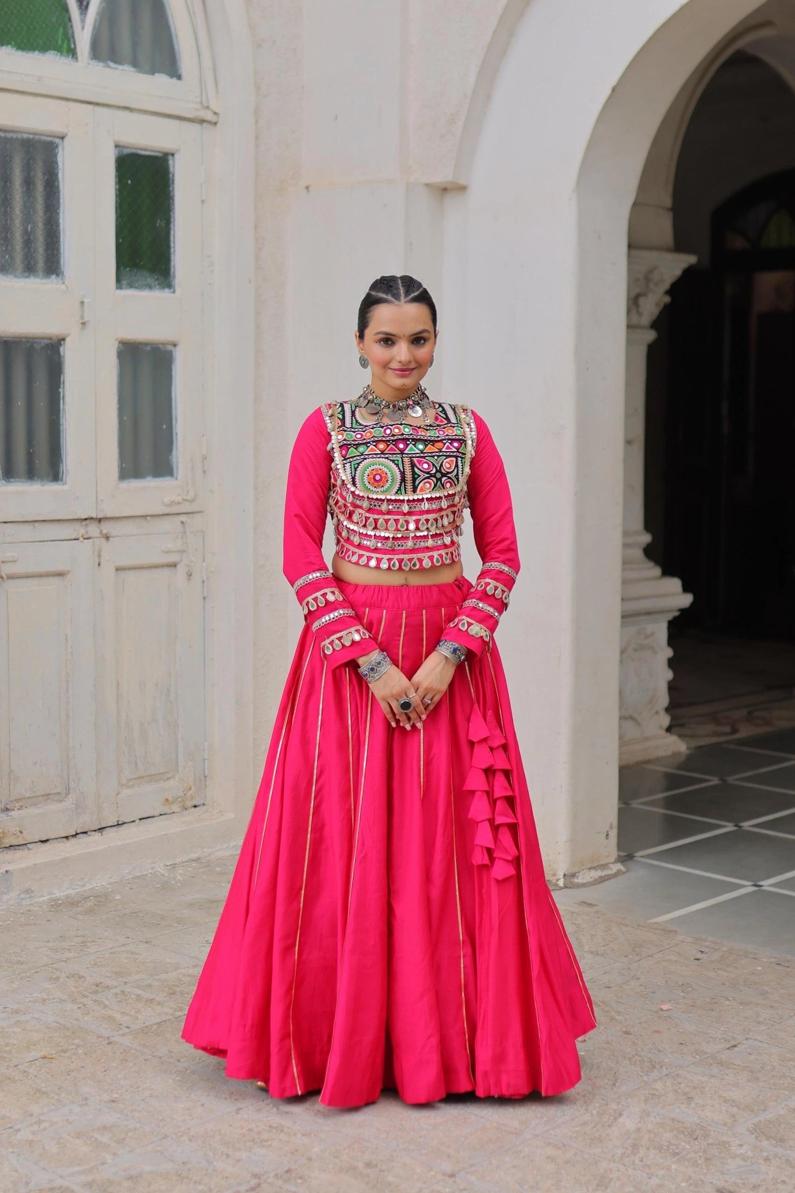 Woman in a traditional pink outfit with intricate designs standing in front of a white building.