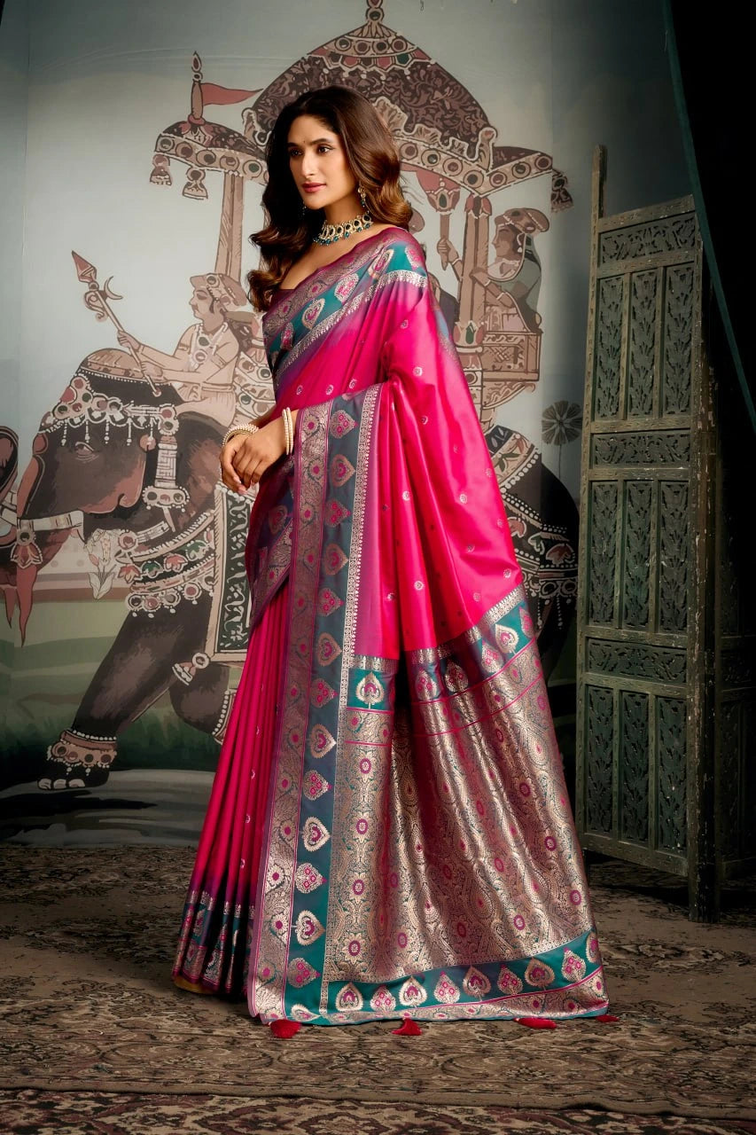 Woman in a pink saree with intricate patterns standing in front of an elephant and architectural background.