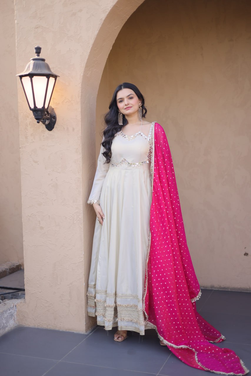 Woman in a white traditional outfit with a pink dupatta standing against a beige wall.