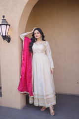 Woman in a white traditional outfit with a pink dupatta standing against a beige wall.