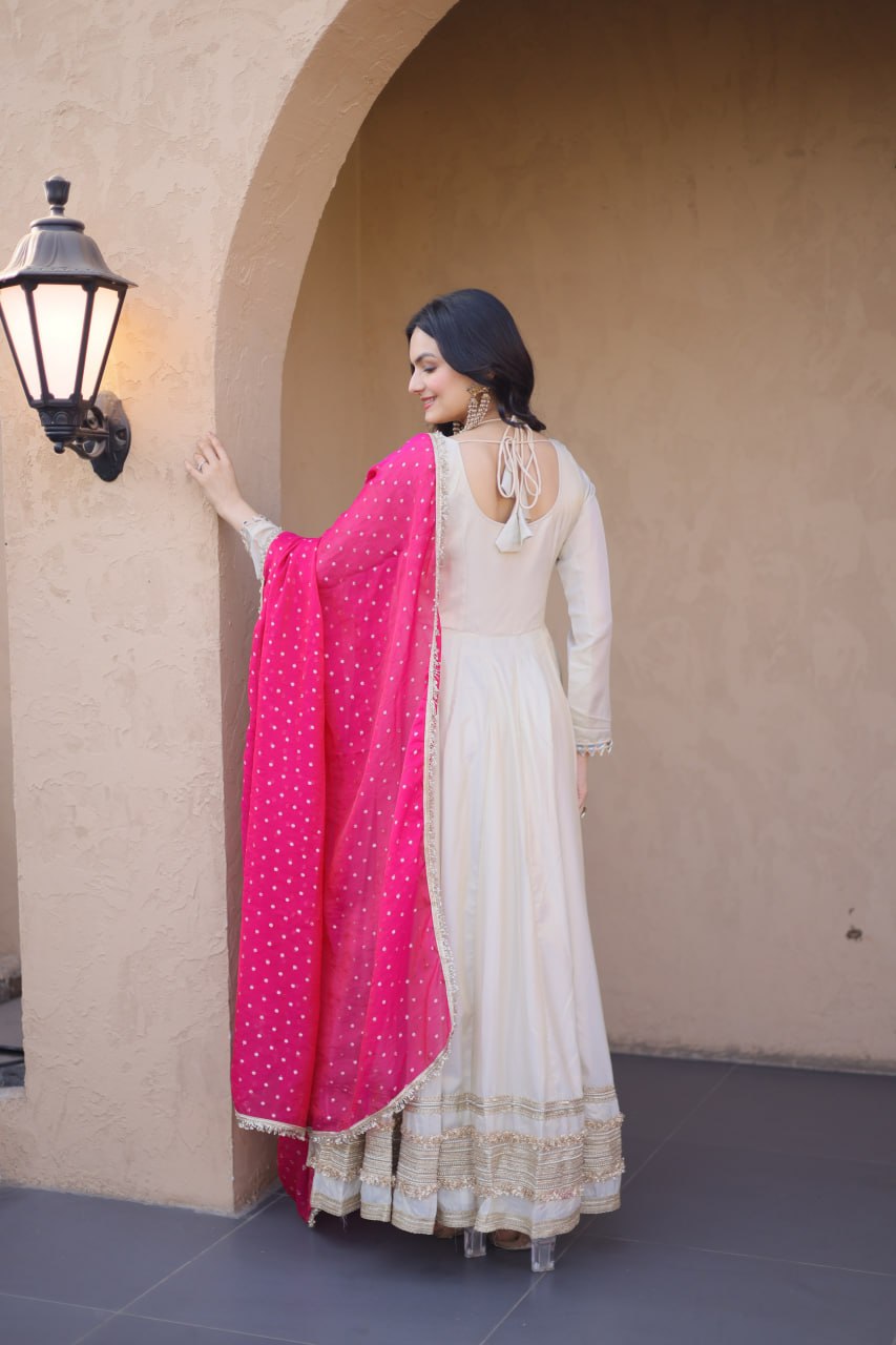 Woman in a white traditional outfit with a pink dupatta standing against a beige wall.