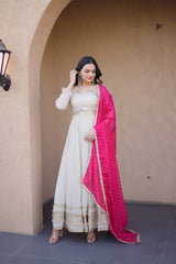 Woman in a white traditional outfit with a pink dupatta standing against a beige wall.