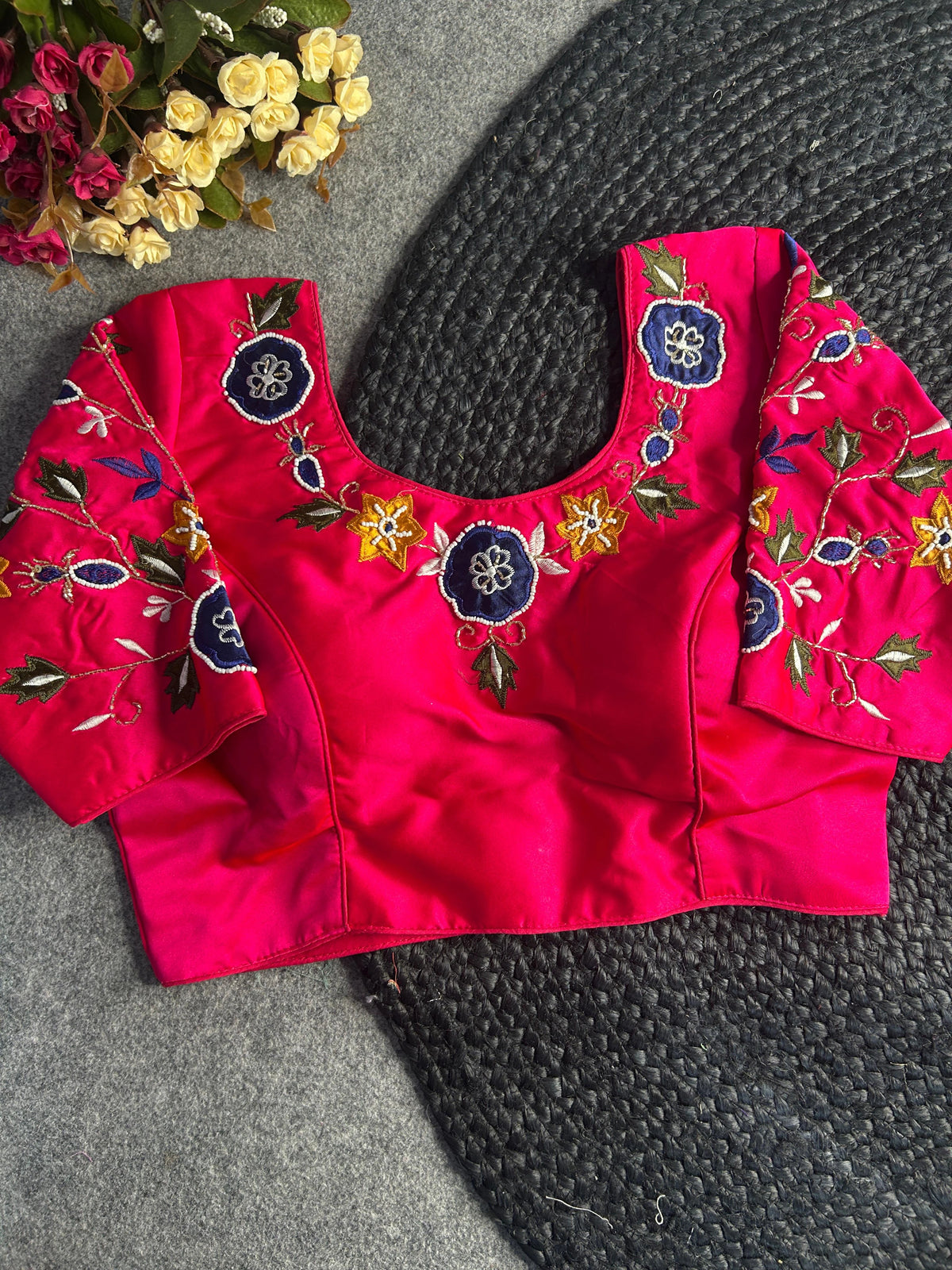 A vibrant pink silk blouse is shown from a side-angled view, highlighting the intricate hand-embroidery on the sleeve and neckline. The design features navy blue and yellow floral motifs framed by olive green and white leaves. Delicate gold thread work connects the floral elements in a vine-like pattern across the lustrous fabric.