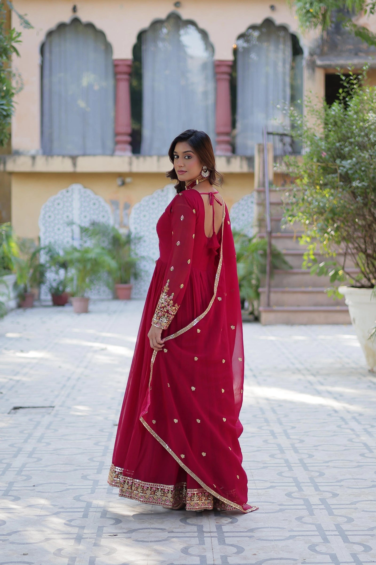 Full-length back view of a woman in a flowing magenta gown. The dress has a deep round back with tie-up detailing. She drapes a matching sheer dupatta over her shoulder in a tiled courtyard with traditional arches.
