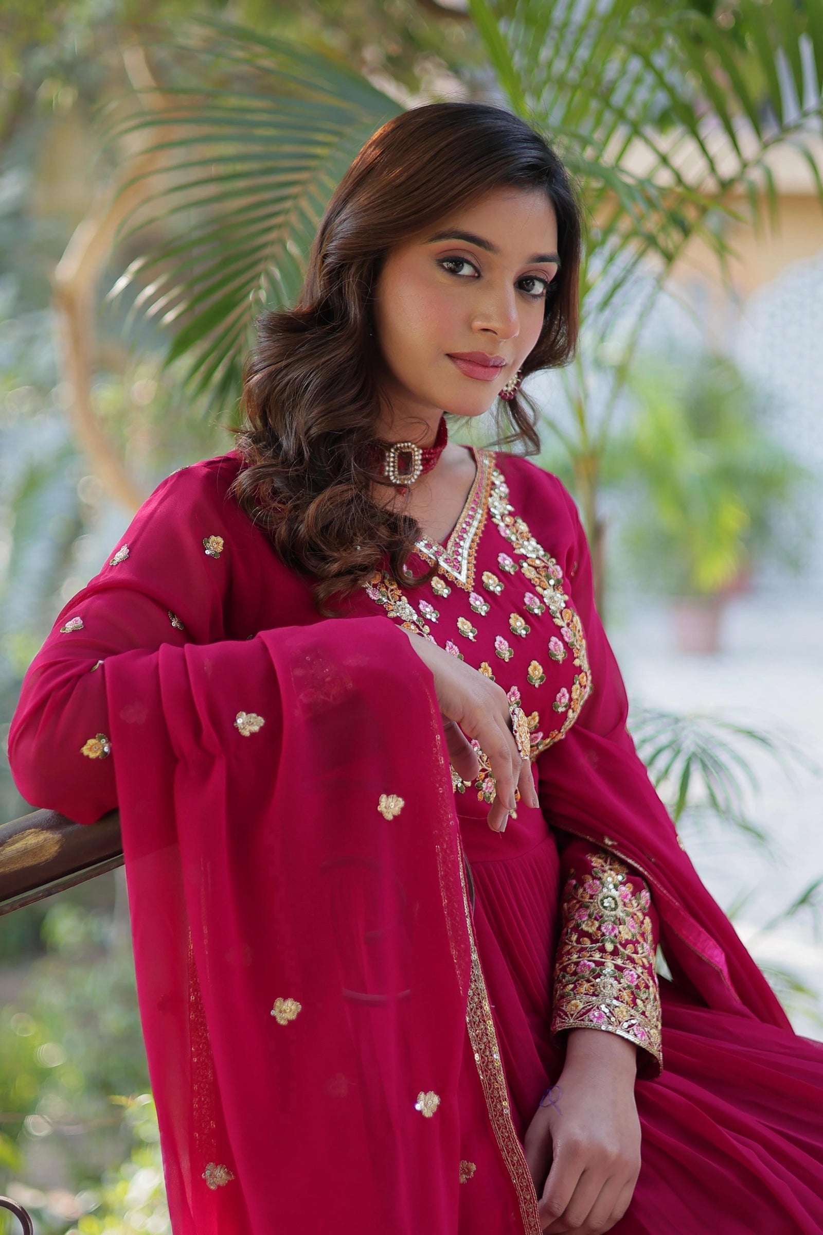 Close-up of a woman in a magenta Anarkali suit. It features a gold and floral embroidered neckline and sleeves. She wears a matching red choker and has wavy brown hair, posing outdoors against green foliage.