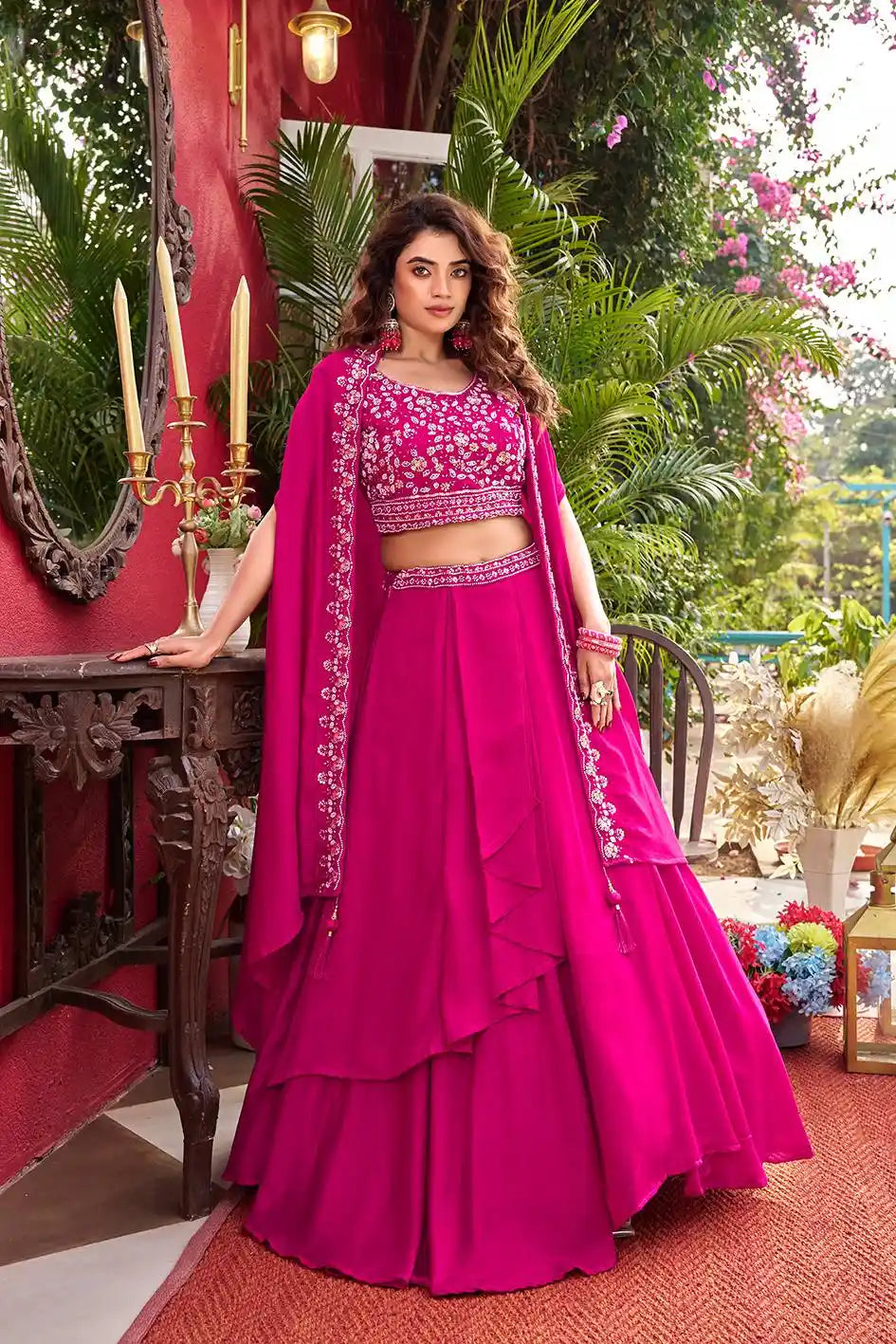 Woman in a vibrant pink traditional outfit with intricate designs, standing in an outdoor setting with greenery.