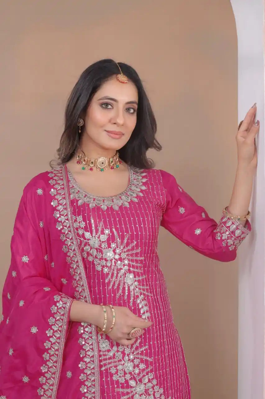 Woman wearing a pink traditional outfit with intricate designs, standing against a beige wall.
