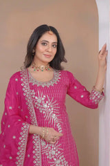 Woman wearing a pink traditional outfit with intricate designs, standing against a beige wall.